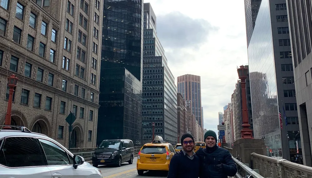 Two individuals are standing on a city bridge with urban architecture and cars around them smiling for the camera