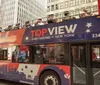 Passengers on an open-top double-decker tour bus are smiling and enjoying the sights of a city street