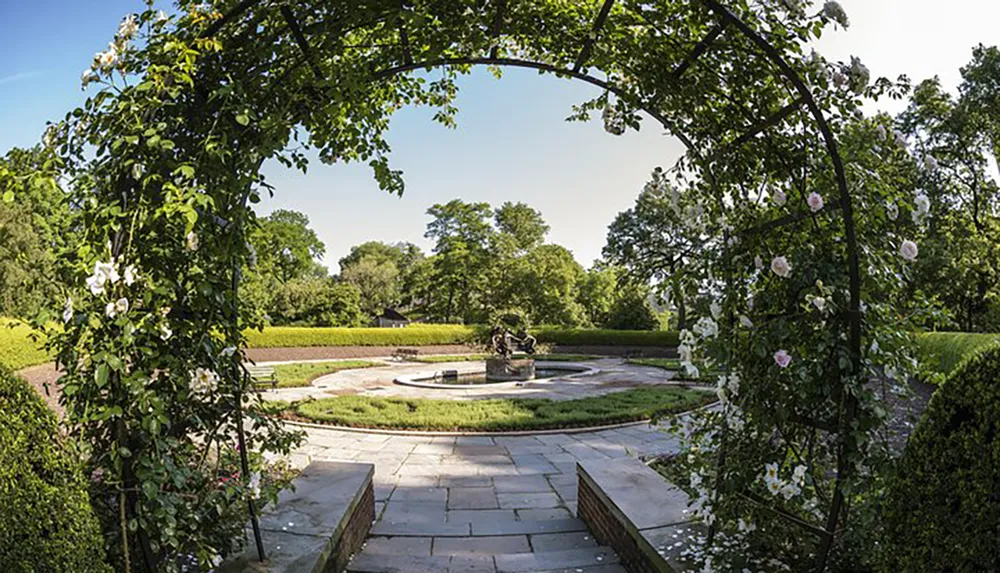 An arched trellis adorned with climbing roses frames a view of a peaceful garden featuring a central sculptural fountain geometric hedge designs and surrounding greenery under a clear sky