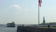 An American flag flies in the foreground with the Statue of Liberty in the distance, while a ferry carries passengers nearby.