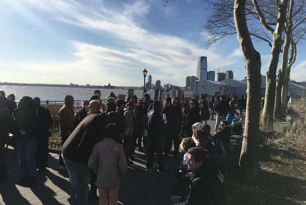 A group of people queueing outdoors on a sunny day with a view of the water and a city skyline in the distance