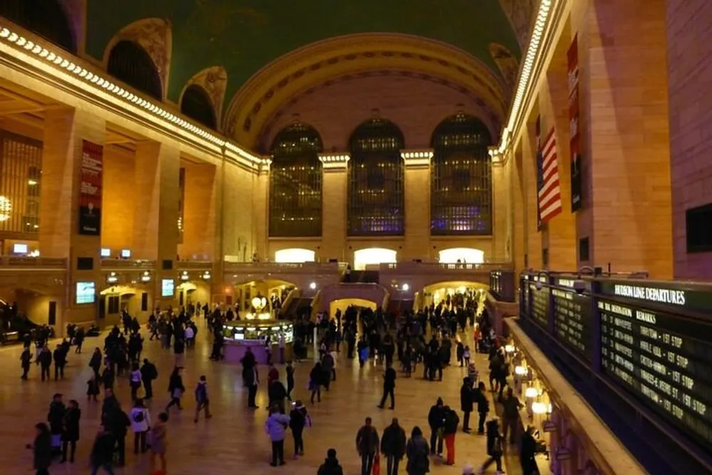 The image shows the bustling interior of a grand train station with high arched windows an American flag and a departure board visible amongst the many passengers