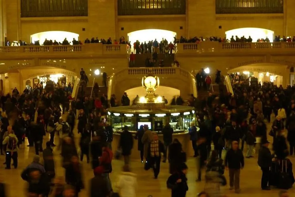 The image shows a bustling scene inside a grand historic train station with people gathered around a central information kiosk below a grand clock with staircases and balconies filled with more individuals