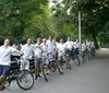 A group of people wearing matching white shirts and gray shorts are standing with bicycles along a park path with some looking at the camera