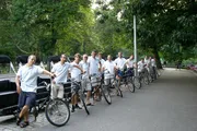 A group of people wearing matching white shirts and gray shorts are standing with bicycles along a park path, with some looking at the camera.