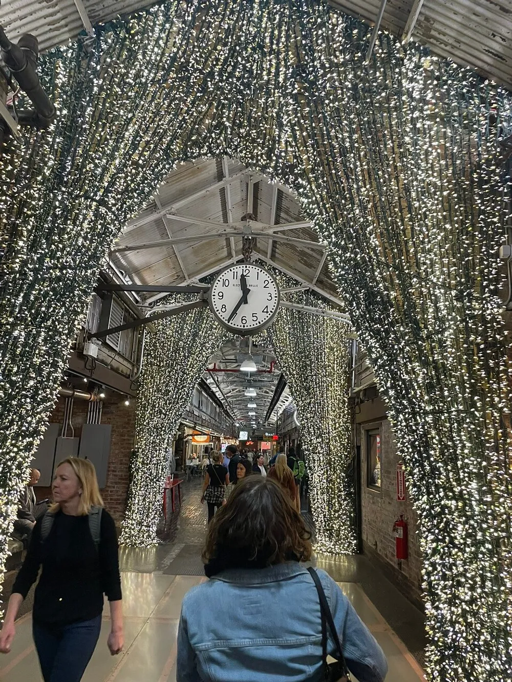 A bustling indoor corridor is adorned with a dazzling canopy of twinkling lights beneath which people stroll past a prominent hanging clock displaying the time