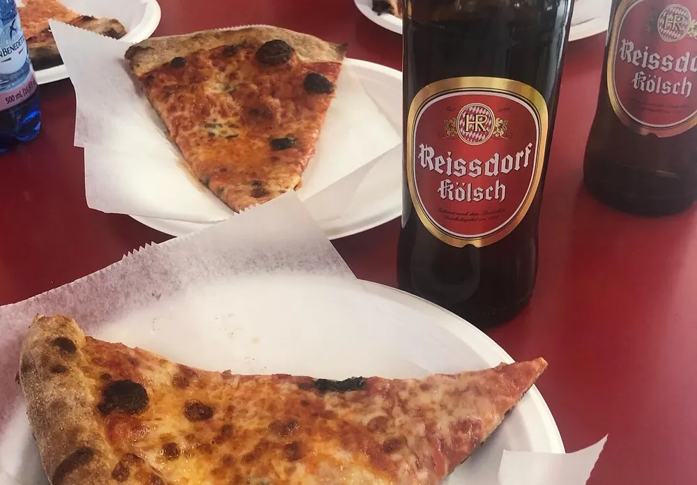 The image shows two slices of pizza on white paper plates alongside two bottles of Reissdorf Klsch beer on a red table