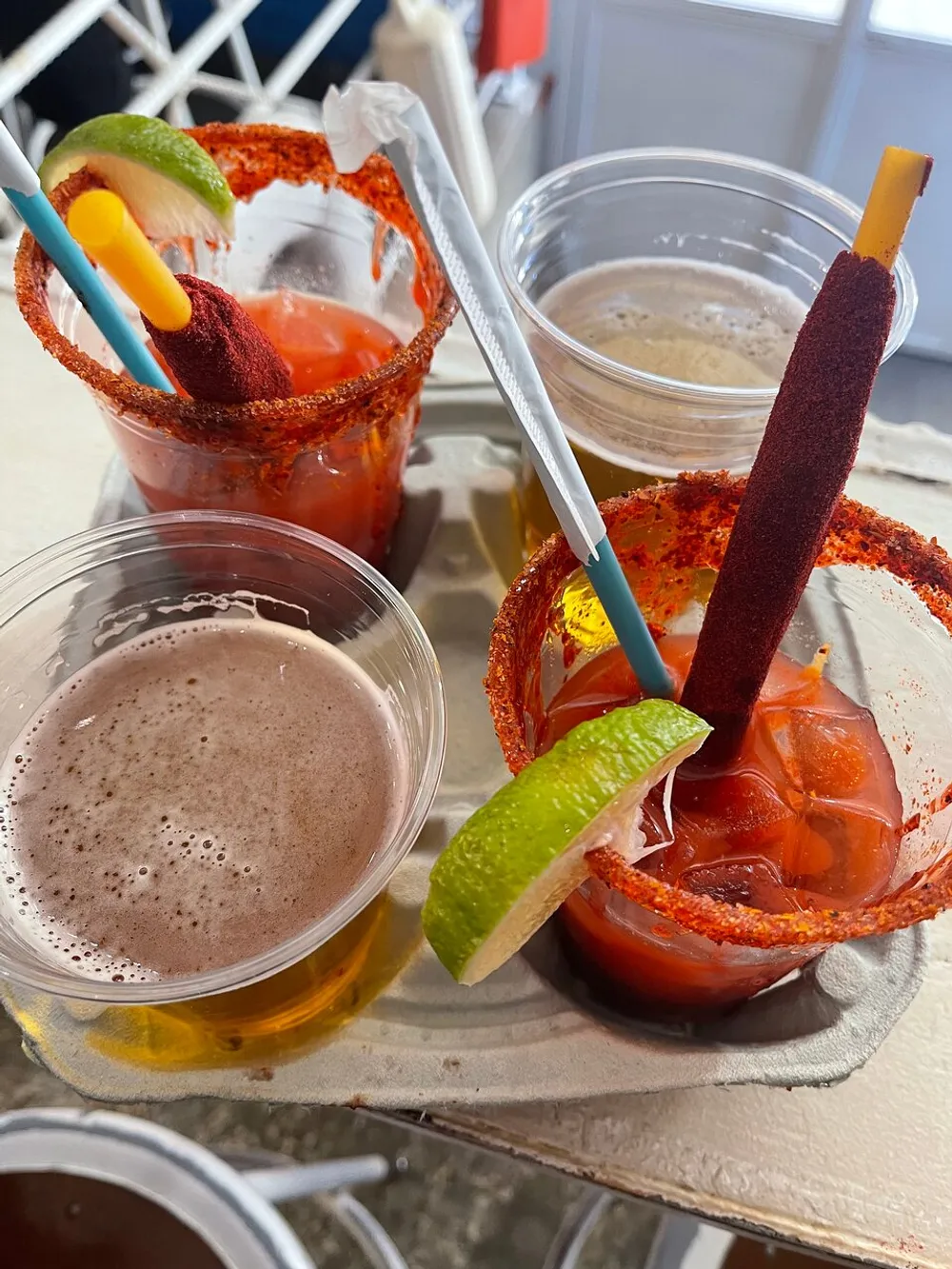 The image shows an assortment of drinks including what appears to be a bloody mary and beers garnished with lime and dressed with a spice rim and snack sticks served on a tray