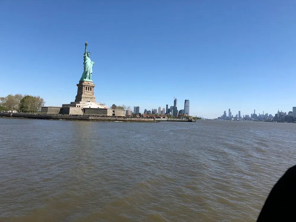 The image shows the Statue of Liberty on its island with a backdrop of a blue sky and the New York City skyline in the distance as viewed from the water
