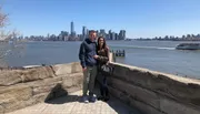Two people are smiling for a photo with the New York City skyline in the background on a clear day.