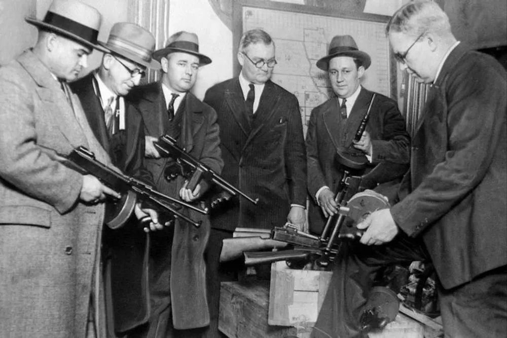 A group of men in suits and hats from an earlier era are examining or displaying a collection of Thompson submachine guns