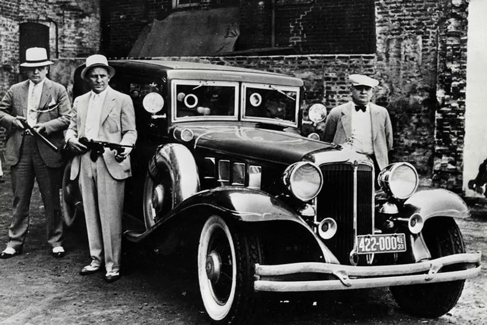 Three men in vintage clothing two wielding firearms stand beside a classic car with a 1930s-style design in front of a brick building
