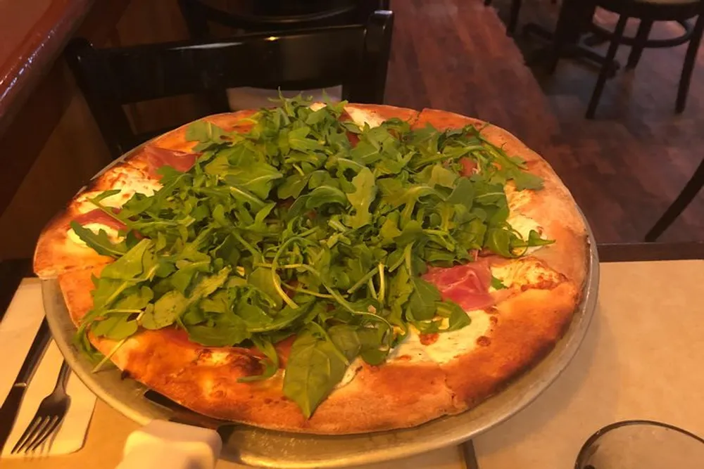 A pizza topped with fresh arugula and slices of prosciutto is presented on a metal stand on a table in a restaurant