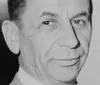 The image shows a close-up of a smiling gentleman with a part of his face obscured presented in black and white