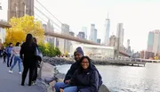 Two people are smiling for a photo in front of the Brooklyn Bridge with the Manhattan skyline in the background.