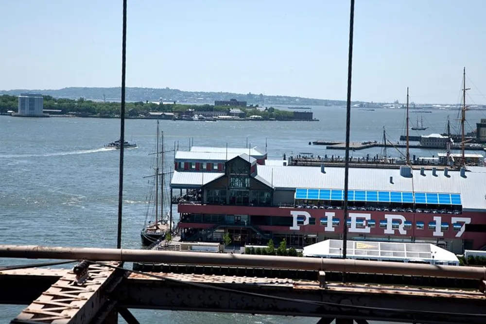 The image shows a waterfront view of Pier 17 with a ship docked nearby taken from above and framed by cables likely from a bridge