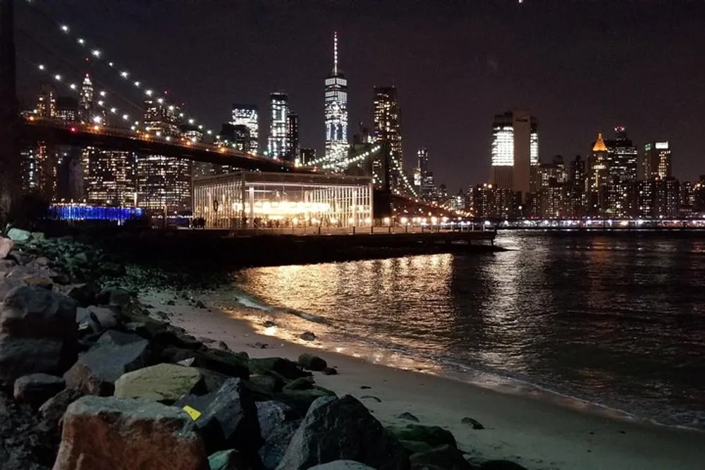 The image shows a night view of a lit-up city skyline with the Brooklyn Bridge on the left overlooking the East River with a small beach area in the foreground