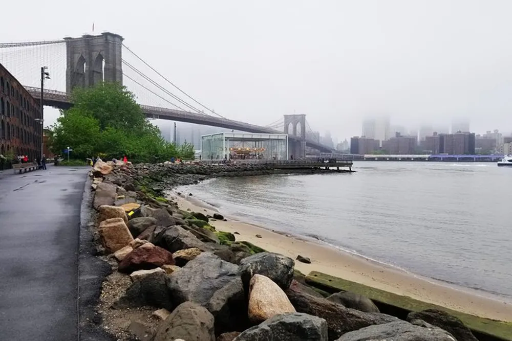 The image features the Brooklyn Bridge on a foggy day with a view from the riverbank showcasing a carousel structure and the mist-obscured skyline of Manhattan in the background