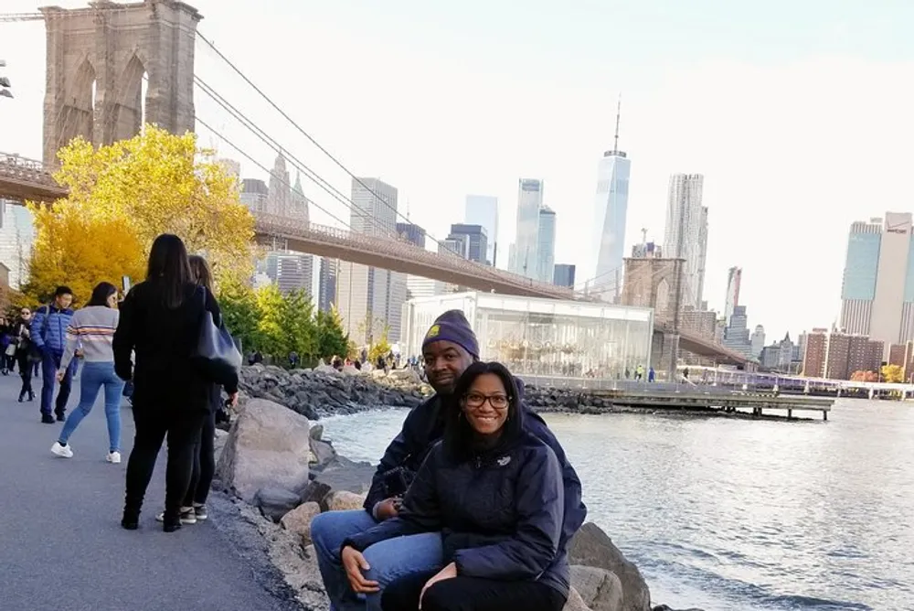 Two people are smiling for the camera while seated on rocks near a waterfront park with the Brooklyn Bridge and the Manhattan skyline in the background