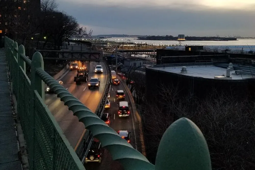 Vehicles are driving on a highway at dusk with a calm river and distant lights in the background viewed from above through a green fence