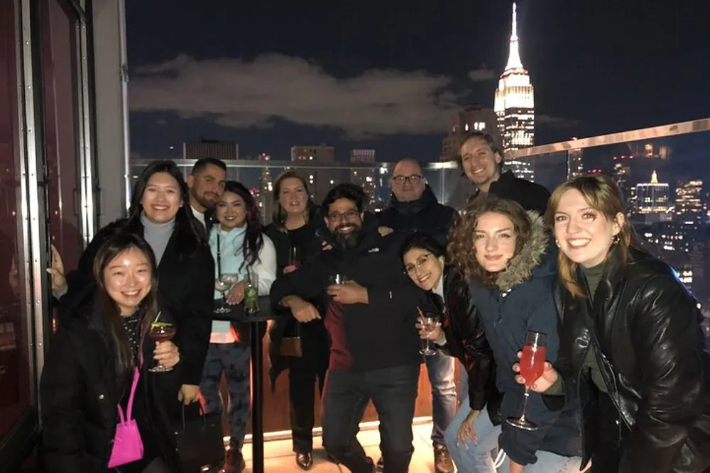 A group of people are posing for a photo at night with drinks in their hands on a rooftop overlooking a city skyline that includes the Empire State Building