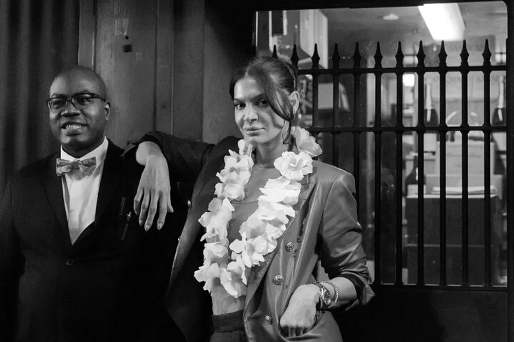 A man in a tuxedo with a bow tie and glasses stands next to a woman wearing a lei and a blazer both of whom are posing with confidence in a monochrome photo