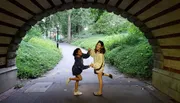 Two people are sharing a playful moment under a brick archway in a lush green park.