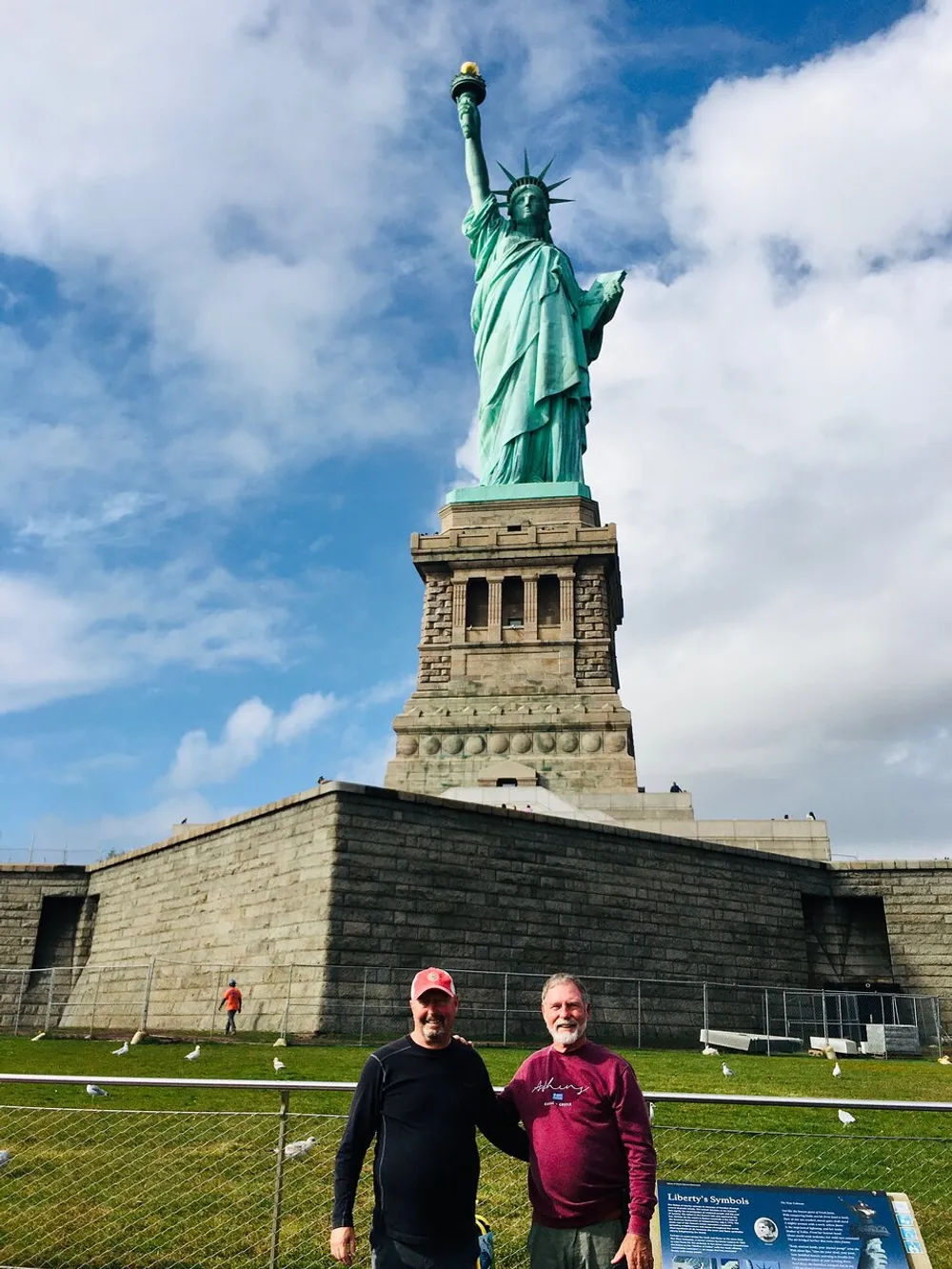 Two individuals are posing for a photo in front of the Statue of Liberty on a day with a partly cloudy sky