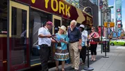 A tour guide is assisting tourists with a map beside a 'Big Bus New York' tour bus, with Times Square in the background.