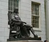 The image shows a bronze statue of a seated figure with a book on their lap set against the backdrop of a building with large windows