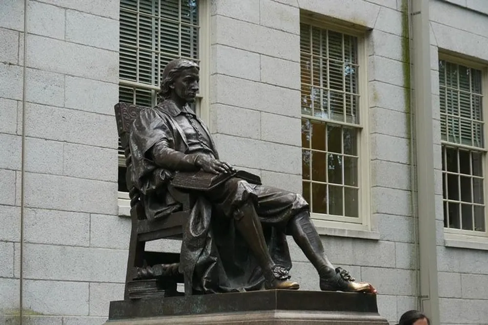The image shows a bronze statue of a seated figure with a book on their lap set against the backdrop of a building with large windows