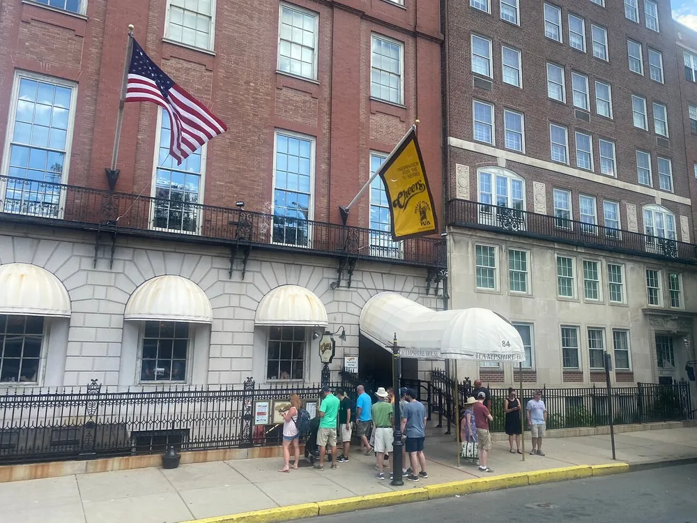 A group of people is queuing outside a building flying the American flag and a yellow banner with a striped awning over the entrance