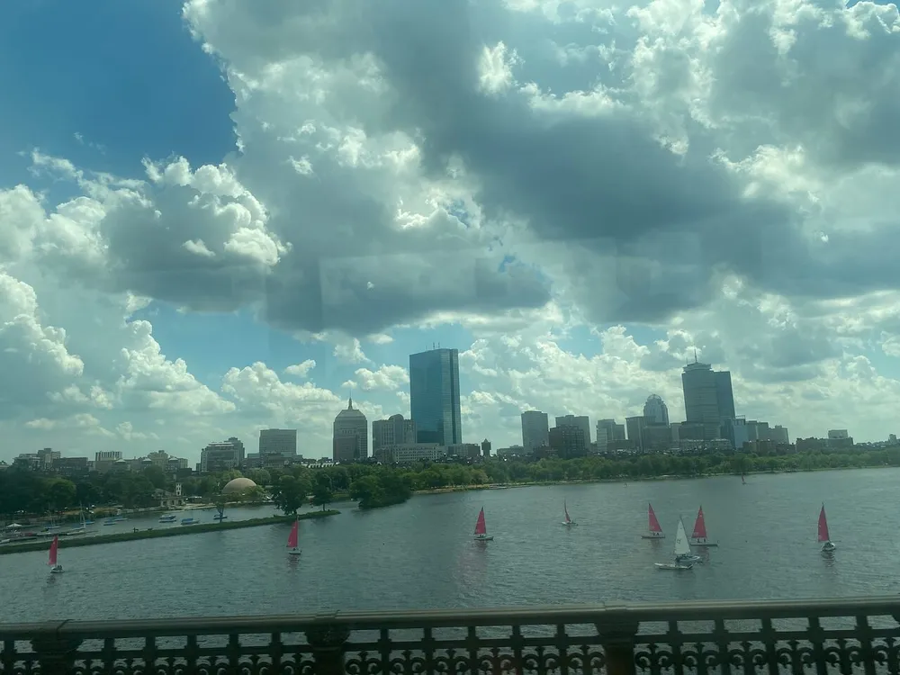 The image shows a view of a city skyline across a body of water with sailboats and a cloudy blue sky as seen through a window with reflections