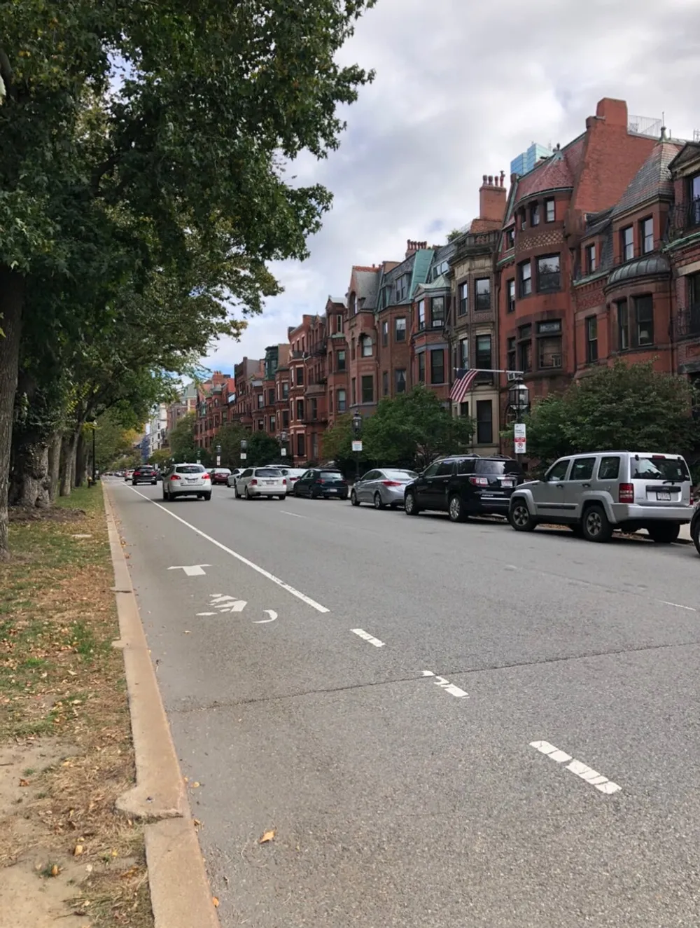 The image shows a tree-lined urban street with a row of parked cars and a series of red brick row houses on what appears to be a cloudy day