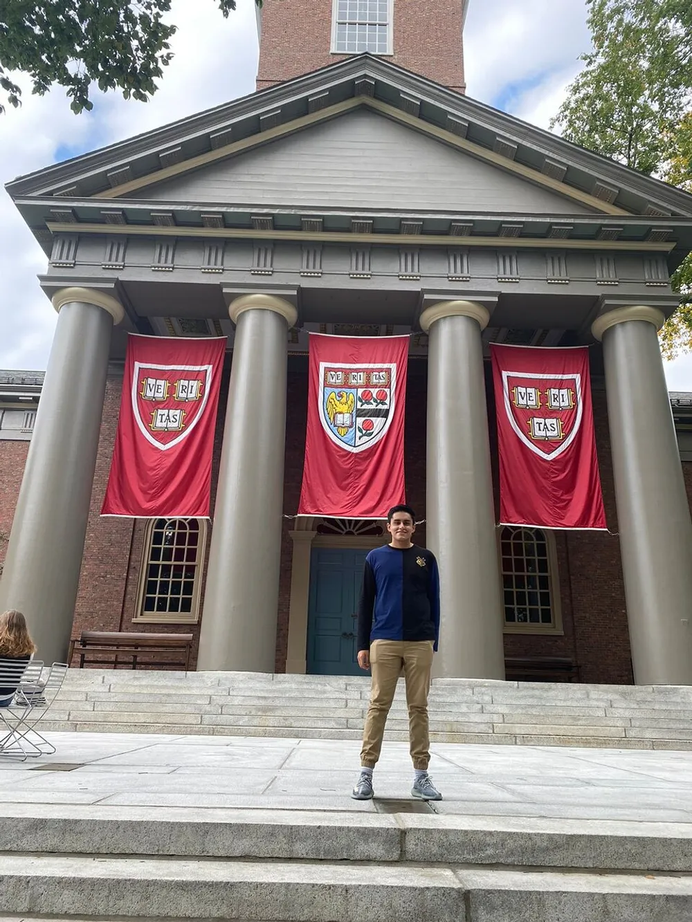 A person stands smiling at the camera in front of a building adorned with crimson banners featuring a shield emblem suggestive of a university setting