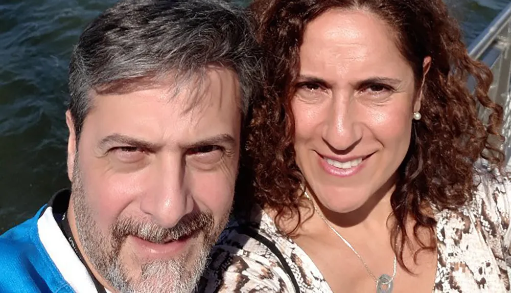 A smiling man and woman are taking a close-up selfie on a sunny day with water and reflections in the background