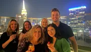A group of people are posing for a photo with drinks at a rooftop venue at night, with the illuminated skyline of a city in the background.