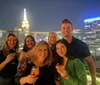 A group of people is posing with drinks on a rooftop at night with a brightly lit city skyline in the background