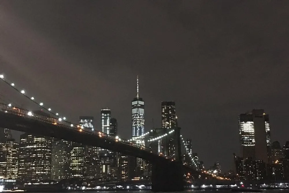 The image captures a night view of a city skyline with illuminated skyscrapers and a notable bridge in the foreground possibly Brooklyn Bridge with the Manhattan skyline in the background