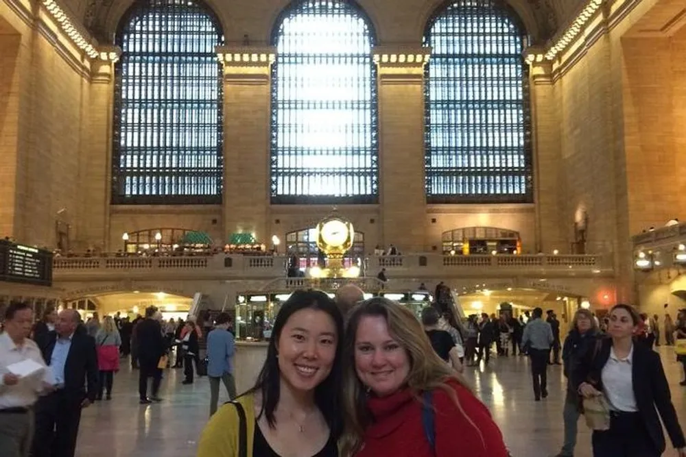 Two smiling women pose for a photo in a busy grand hall with large windows and a clock likely in a historic train station