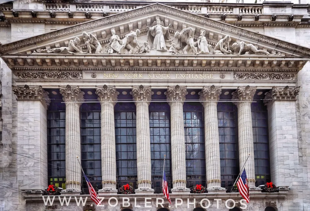 The image shows the neoclassical facade of the New York Stock Exchange with its iconic Corinthian columns and a sculptural pediment accented with American flags and holiday wreaths