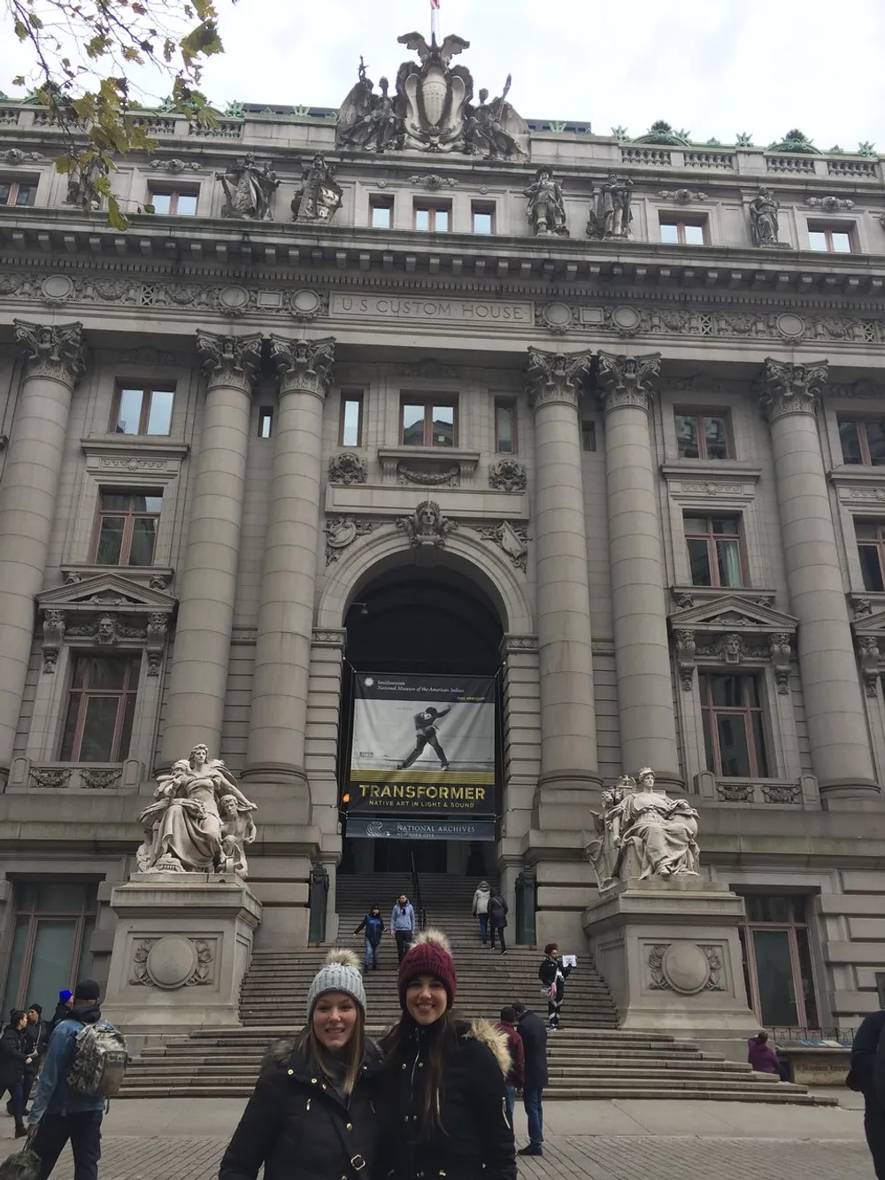 Two people are smiling in front of a grand historical building marked as the US Custom House with a banner promoting a Transformer exhibition at the National Archives