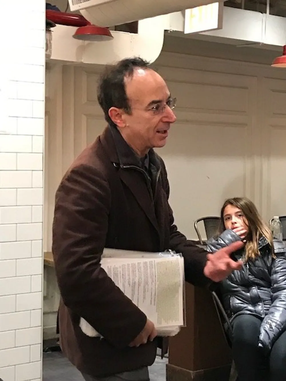 A man in a brown jacket is speaking and gesturing with his right hand while holding papers with a young girl seated in the background