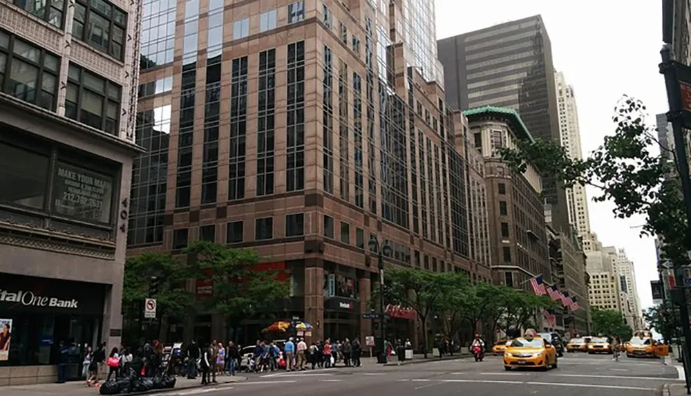 This is a bustling city street lined with tall buildings and featuring pedestrians taxis and American flags fluttering in the wind