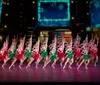 A group of dancers dressed in festive red and green costumes is performing a synchronized routine on a stage with a wintery holiday-themed backdrop