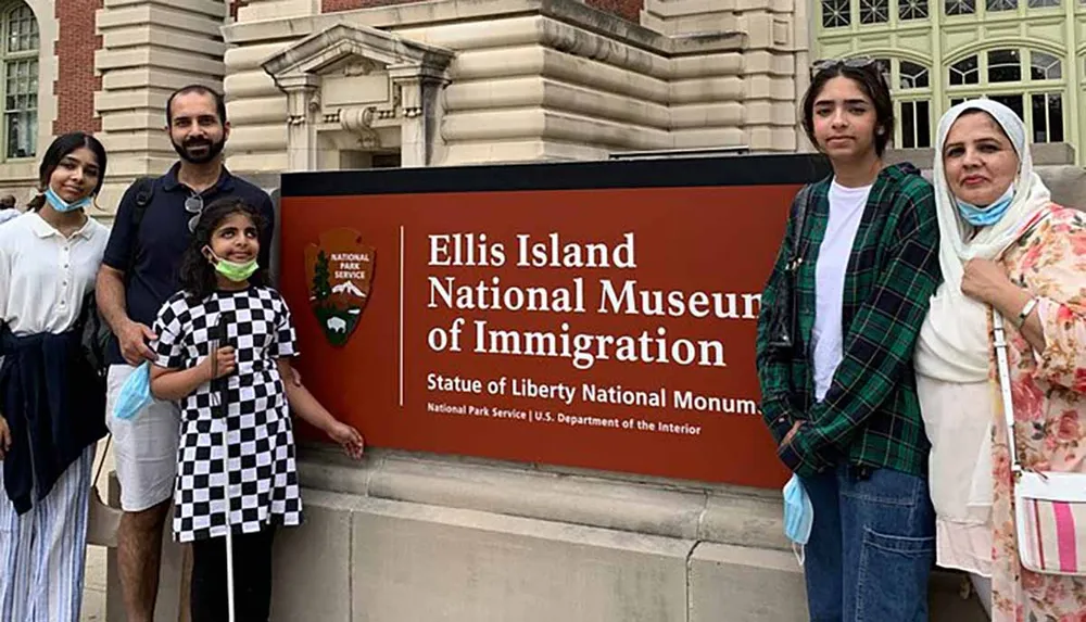 A family of four poses for a photo in front of a sign that reads Ellis Island National Museum of Immigration - Statue of Liberty National Monument