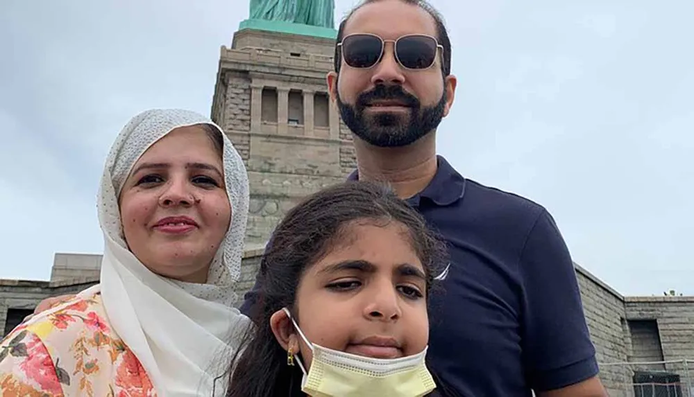 A family of three is posing for a photo with the Statue of Liberty in the background