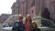 Three people are smiling in front of a fountain with the facade of a grand building in the background on a sunny day.