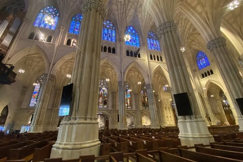 The image shows the interior of a grand gothic-style cathedral with high vaulted ceilings stained glass windows and rows of wooden pews
