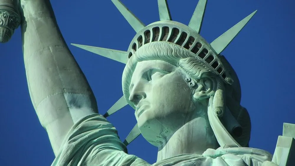 The image shows a close-up of the Statue of Liberty against a clear blue sky highlighting her iconic crown and torch